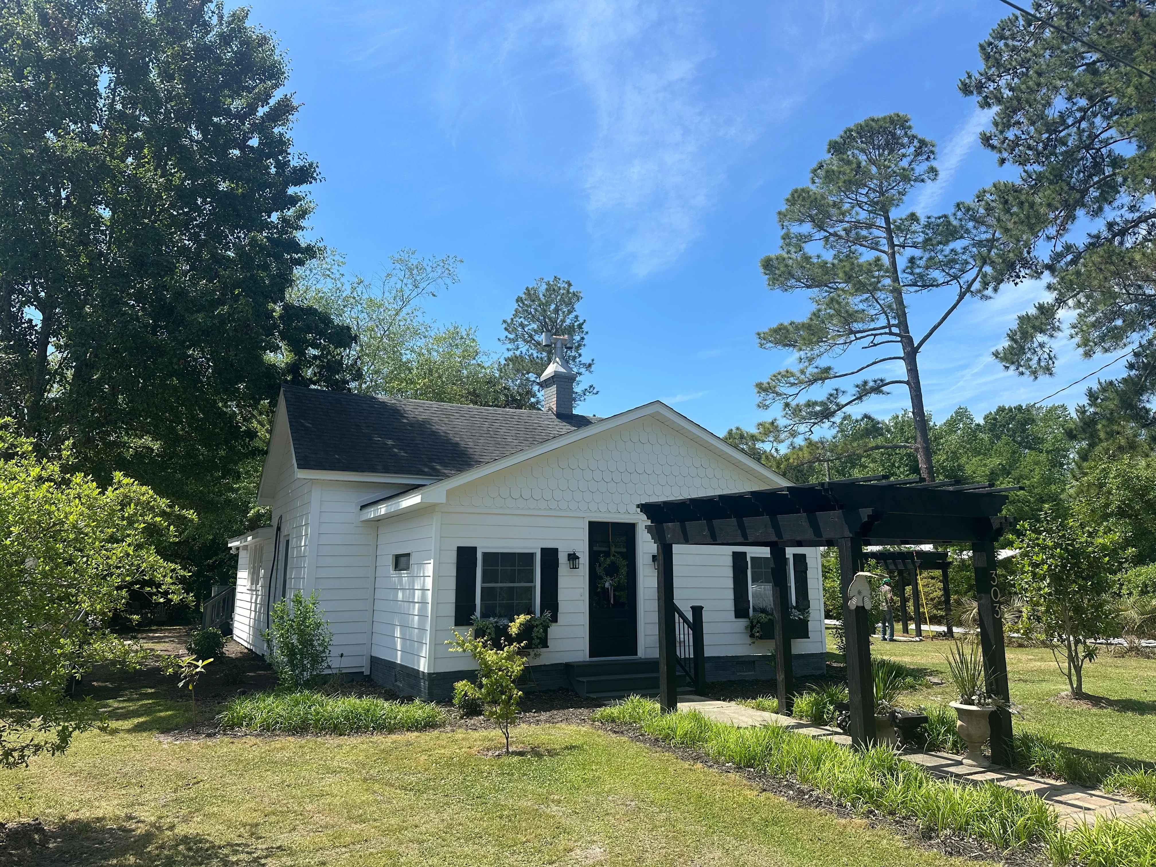 White cottage with dark gray dimensional shingle roof, black shutters, and front porch pergola